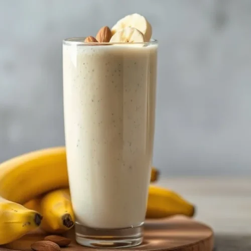 Creamy banana smoothie in a tall glass with sliced bananas, almonds on top, fresh ripe bananas beside, natural daylight, appe